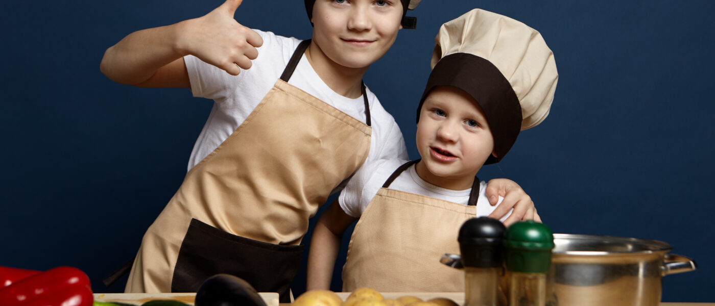 Deux enfants en tenue de petit chef préparent un plat avec des légumes frais dans une cuisine, pour illustrer la Journée internationale des chefs.