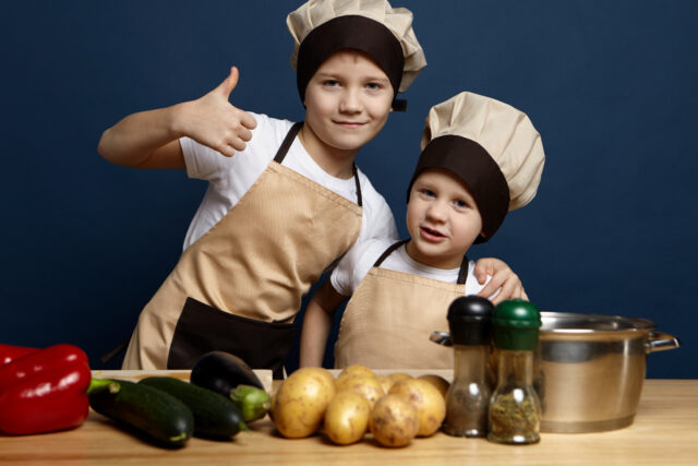 Deux enfants en tenue de petit chef préparent un plat avec des légumes frais dans une cuisine, pour illustrer la Journée internationale des chefs.